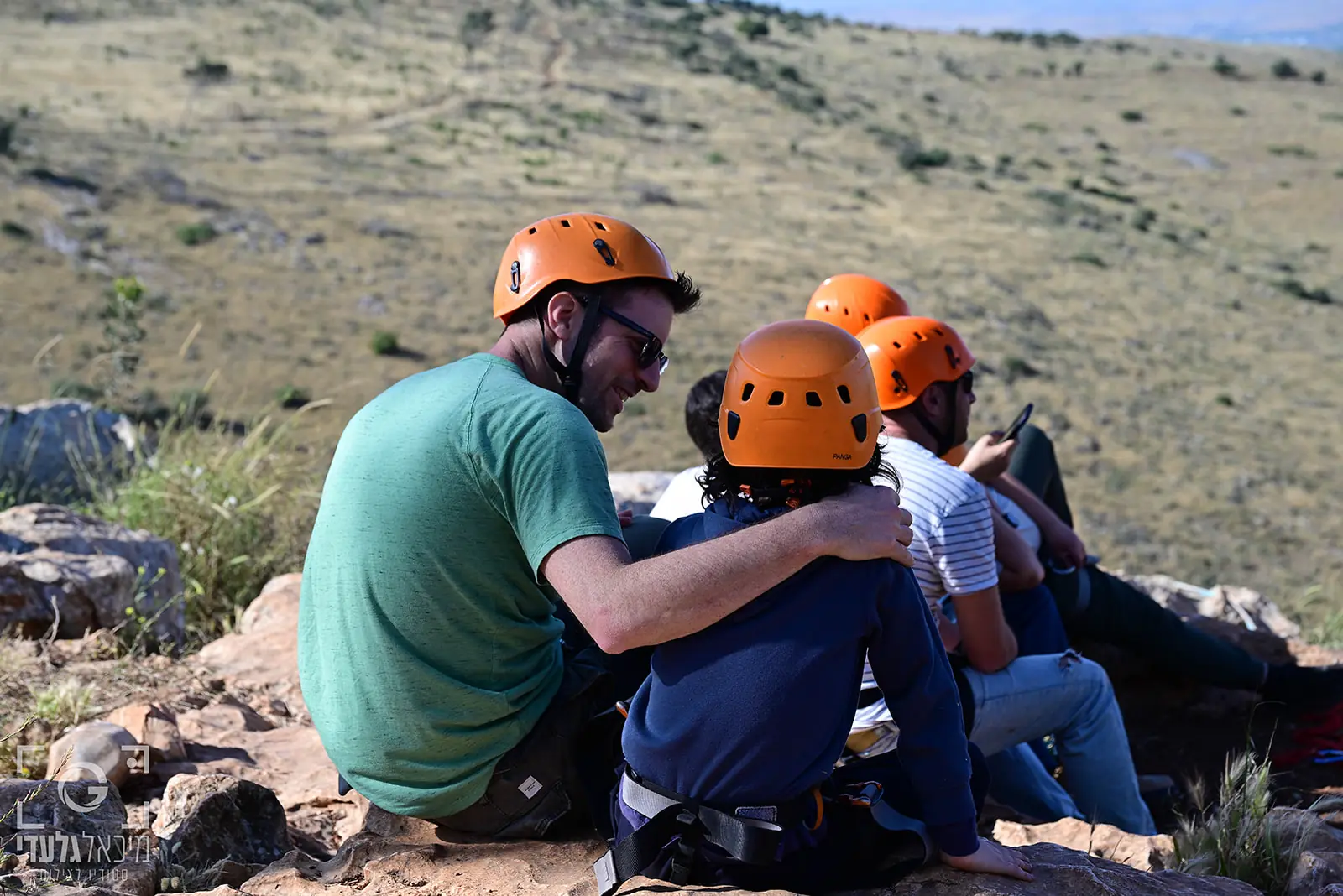 אבא מחבק את בנו, שניהם בקסדות, על סלע מעל עמק פתוח